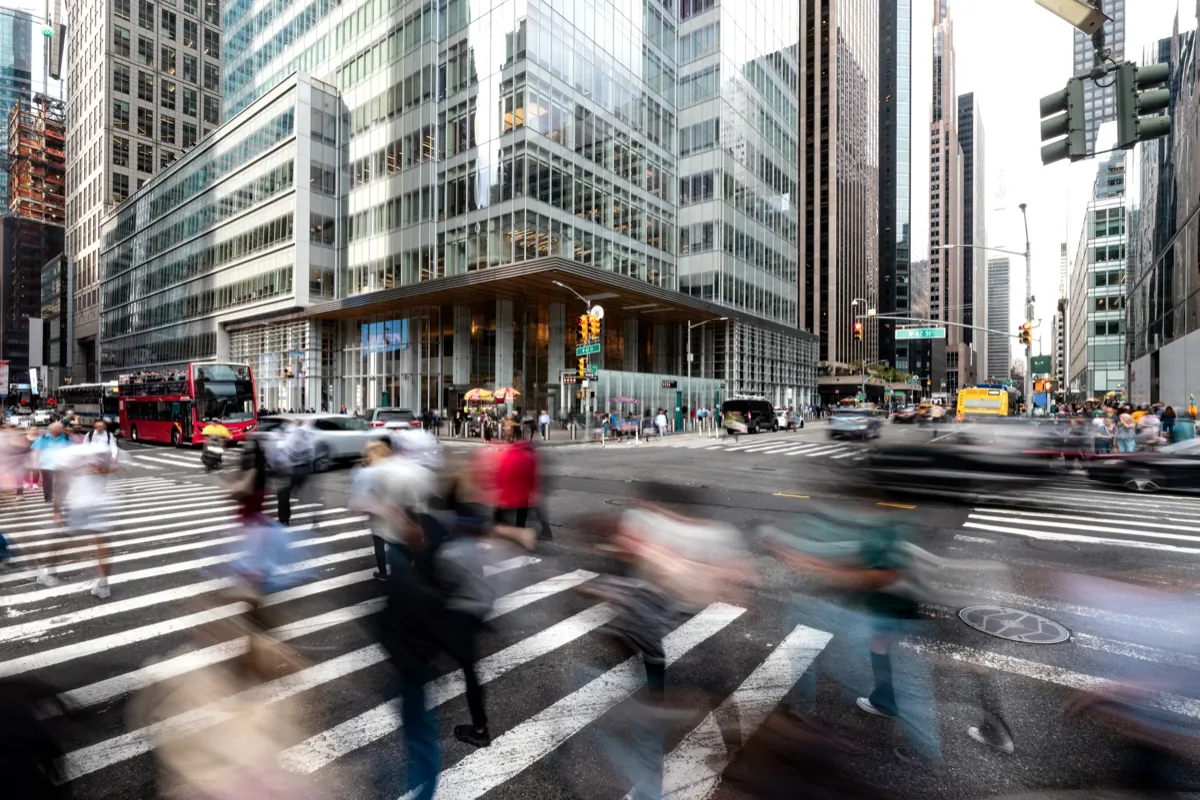Motion-blurred pedestrians crossing a busy New York City intersection surrounded by glass skyscrapers