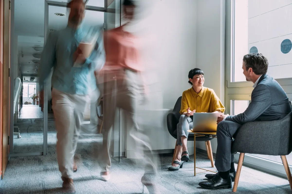 Two colleagues having a seated conversation with laptops in a modern office while blurred figures walk past