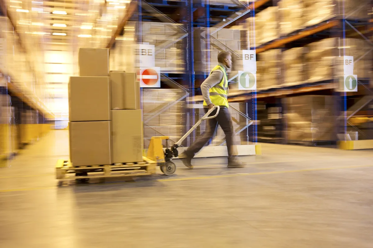 Warehouse worker in high-visibility vest pulling a pallet jack loaded with boxes through a storage facility