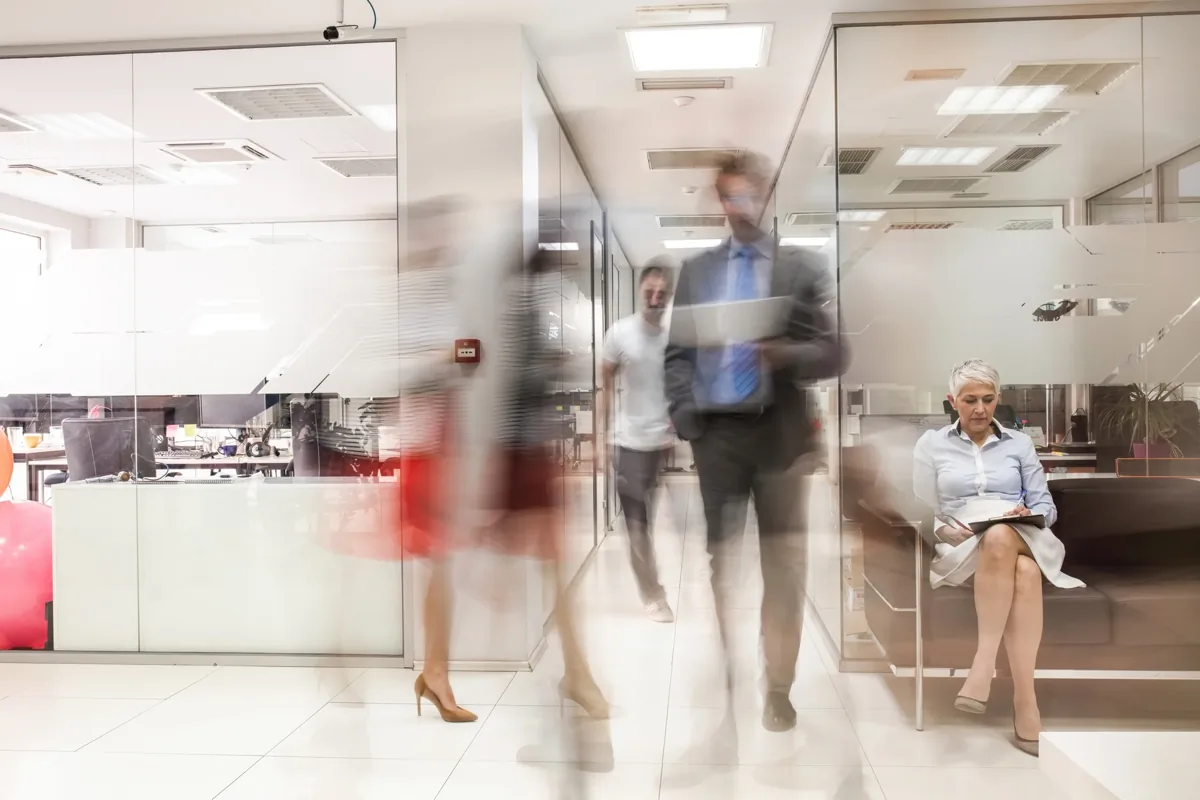 Motion-blurred professionals walking through a modern glass-walled office while a woman reviews documents