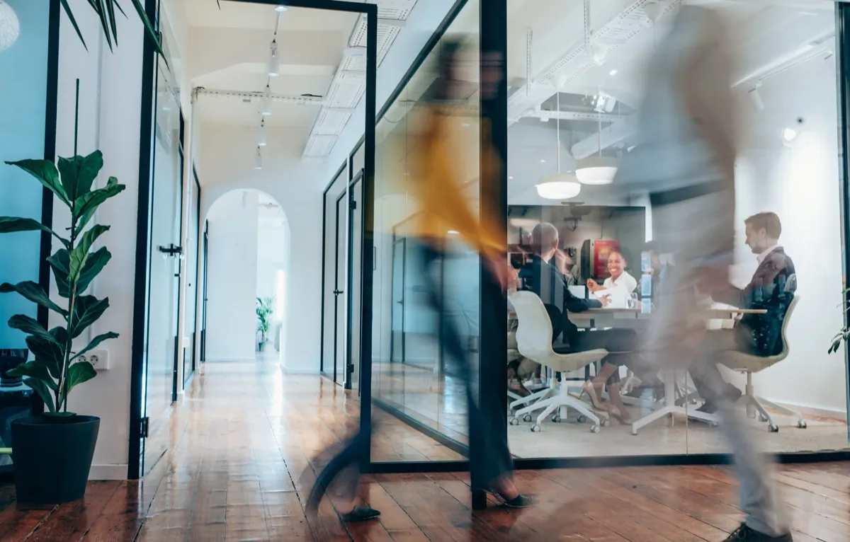 Blurred person walking past a glass-enclosed meeting room where colleagues collaborate in a coworking space