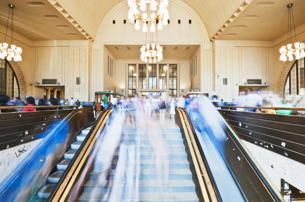 Long-exposure photo of blurred people on escalators and stairs in an ornate transit hall with chandeliers